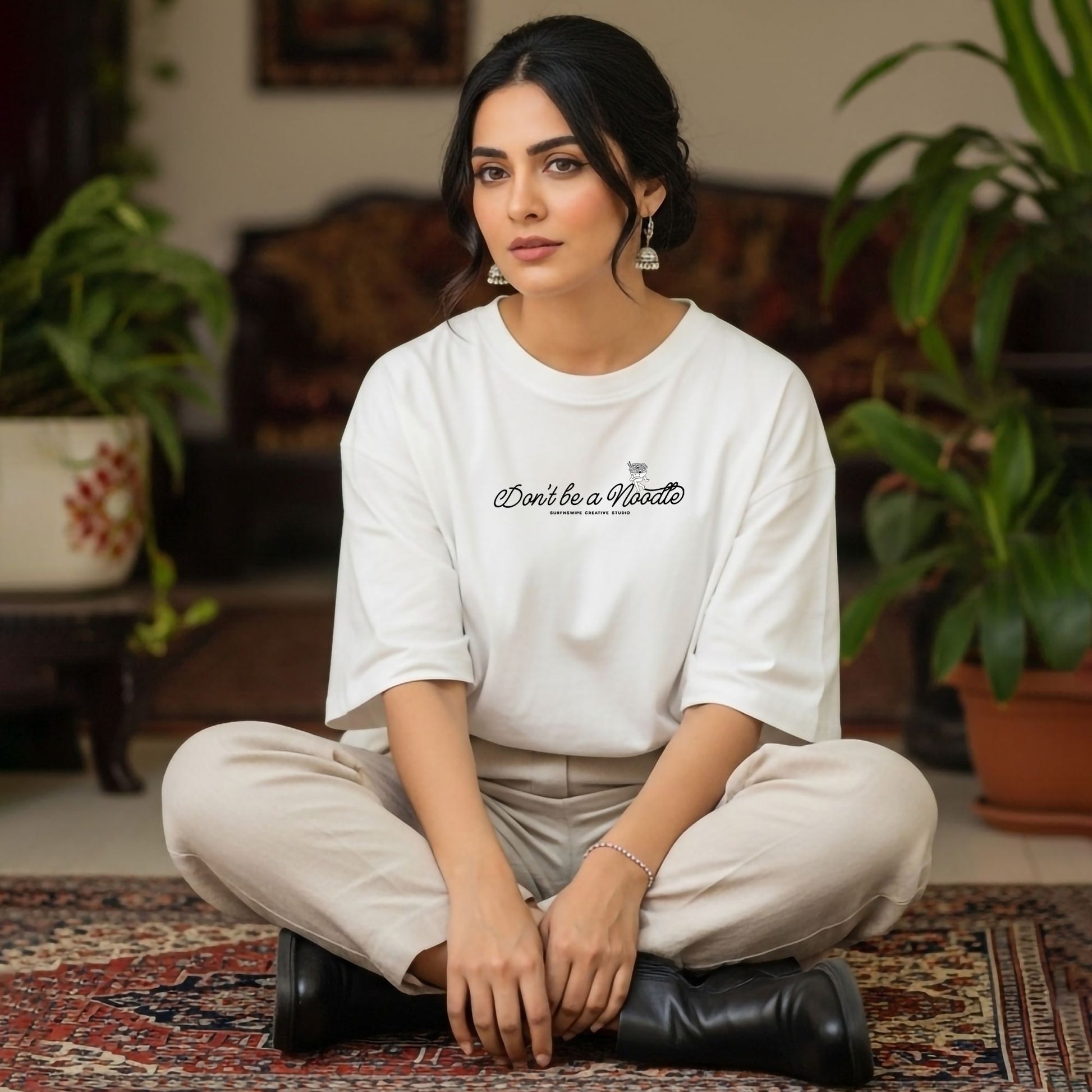 Woman sitting cross-legged on a rug wearing a white t-shirt with text, surrounded by plants.