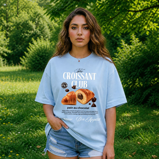 Woman wearing a light blue t-shirt with 'Croissant Club' design outdoors.