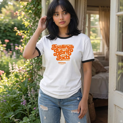 A woman wearing a white ringer T-shirt with black trim and a retro orange graphic that reads 'Feeling Myself One Step At A Time' with 'SURFNSWIPE' below, standing in a sunlit garden doorway next to a bed.