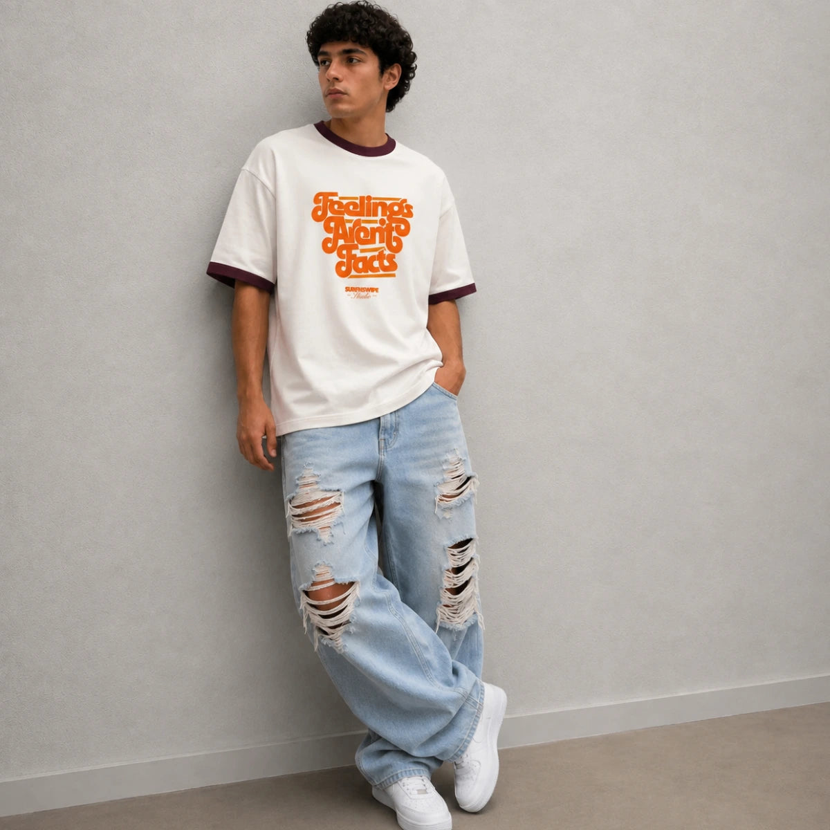 A man wearing a white ringer t-shirt with orange retro text that reads 'Feelings Are Mutually' and light-wash distressed jeans, leaning against a gray wall.
