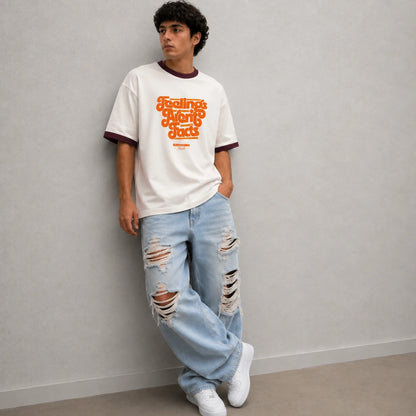 A man wearing a white ringer t-shirt with orange retro text that reads 'Feelings Are Mutually' and light-wash distressed jeans, leaning against a gray wall.