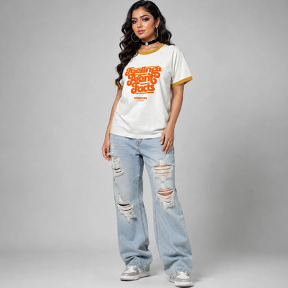 A woman modeling a white ringer t-shirt with orange retro text and yellow trim, paired with distressed light blue jeans and white sneakers, against a gray studio background.