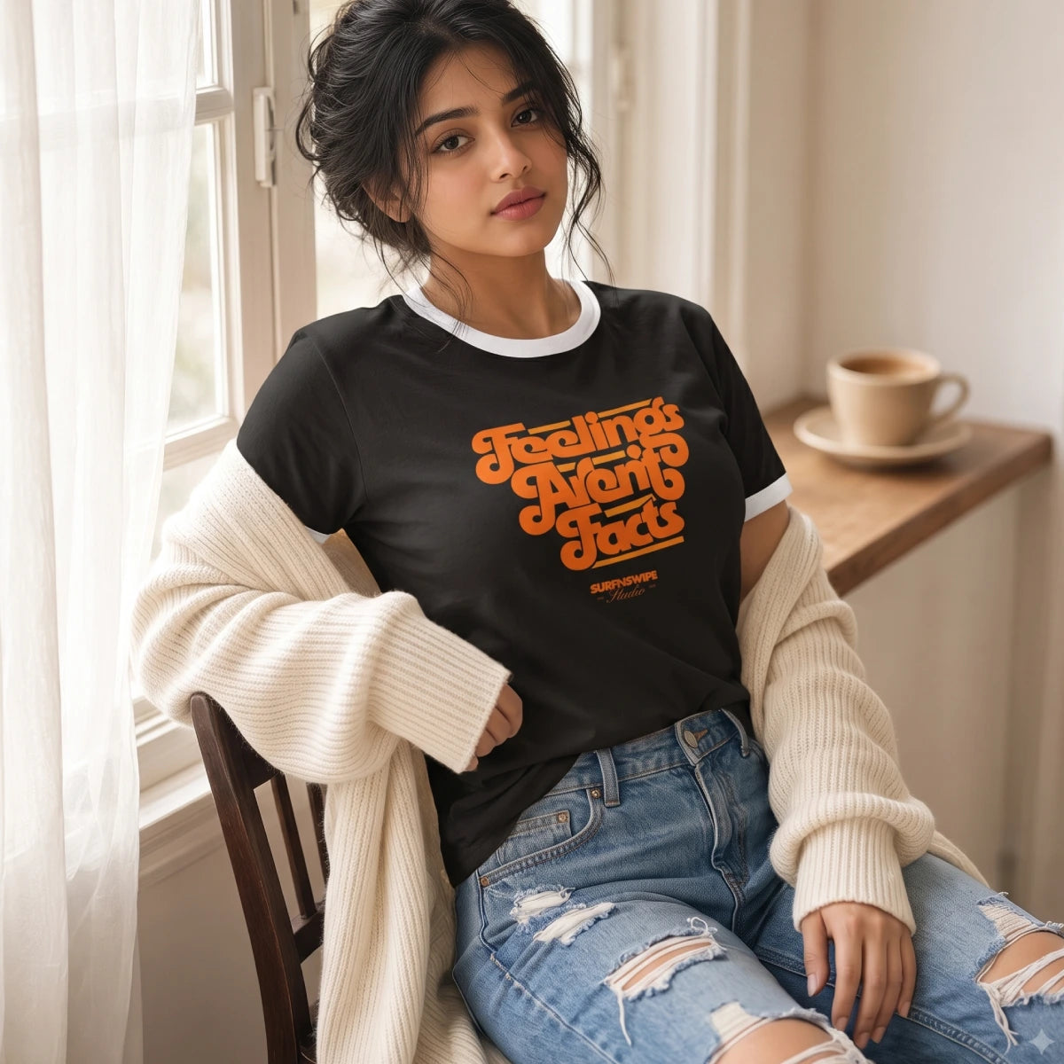 A woman sitting in a chair by a window wearing a black ringer T-shirt with orange retro text reading 'Feeling Myself Again' and a cream ribbed sweater draped off her shoulders, paired with distressed blue jeans.