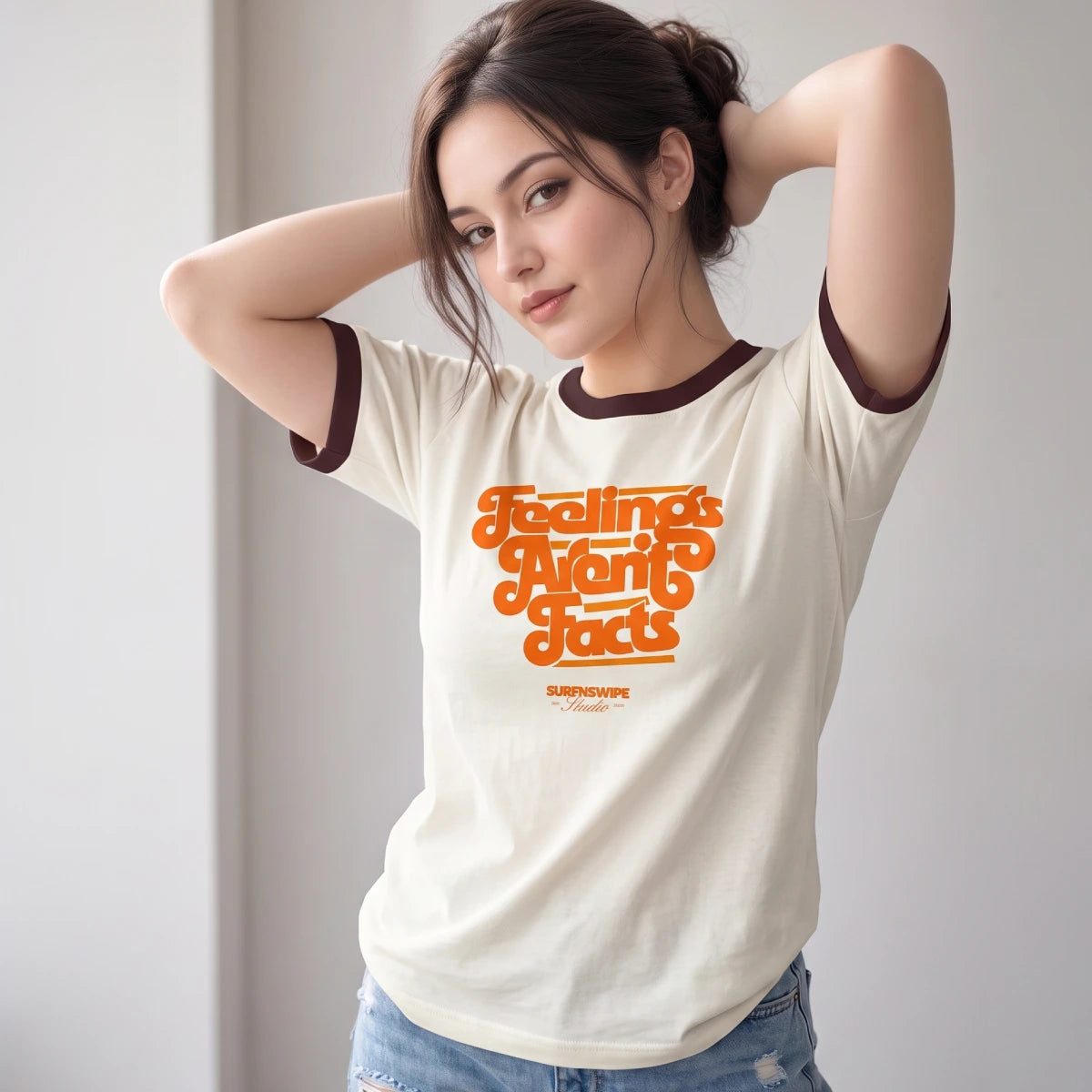 A woman wearing a cream-colored ringer t-shirt with dark brown trim on the collar and cuffs, featuring a retro-style orange graphic that reads 'Feelings Aren't Facts' with 'SURFNSWIPE Collection' printed below. She is posing with her hands behind her head in a brightly lit indoor setting.