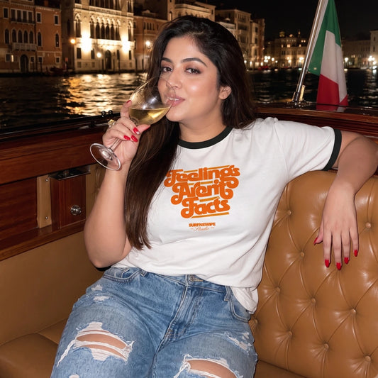 A woman sitting on a tufted brown leather boat seat at night, wearing a white ringer T-shirt with orange retro text that reads 'Feelings Are Facts' and smaller 'SARAHSHI' branding. She is holding a glass of white wine, with a canal and illuminated buildings in the background.