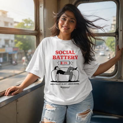 A woman wearing a white graphic t-shirt with a red and black 'Social Battery' design, posing on a bus with city scenery visible through the windows.