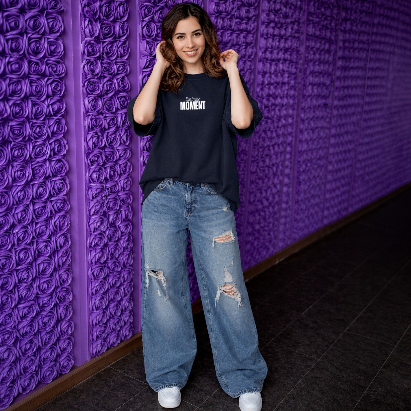 Woman wearing a black t-shirt and blue jeans standing against a textured purple wall.