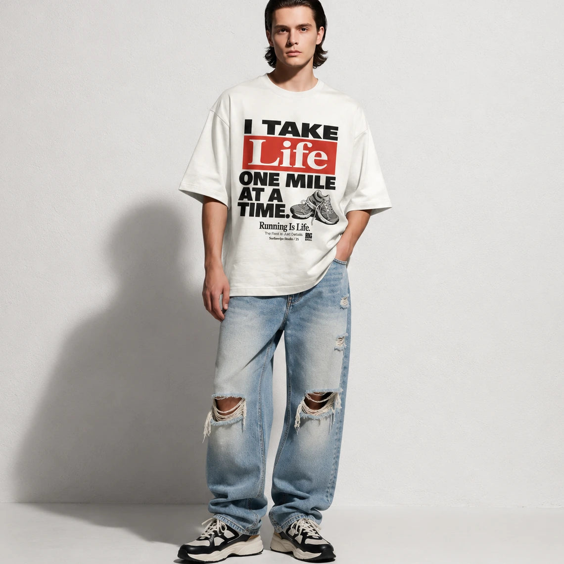 A man stands against a light gray studio wall wearing an oversized white graphic T-shirt with bold black and red text reading 'I TAKE Life ONE MILE AT A TIME,' light blue distressed jeans with knee rips, and black-and-white sneakers.