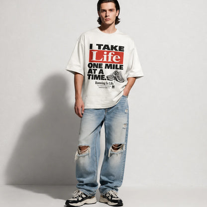 A man stands against a light gray studio wall wearing an oversized white graphic T-shirt with bold black and red text reading 'I TAKE Life ONE MILE AT A TIME,' light blue distressed jeans with knee rips, and black-and-white sneakers.