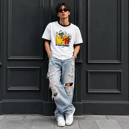 A man wearing a white graphic t-shirt with a colorful cartoon design and the text 'IT IS HAPPENING', paired with distressed light blue jeans and white sneakers, standing in front of a dark paneled door.