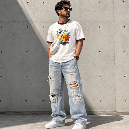 A man wearing a white graphic t-shirt with dark maroon trim, distressed light blue jeans, and white sneakers, standing against a concrete wall.