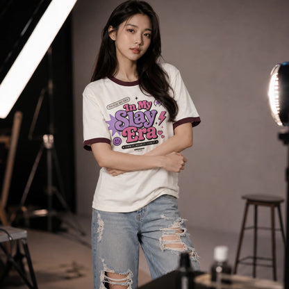 A woman stands in a studio setting wearing a white graphic T-shirt with dark maroon trim and distressed light-wash jeans. The shirt features a colorful front print reading 'In My Slay Era.