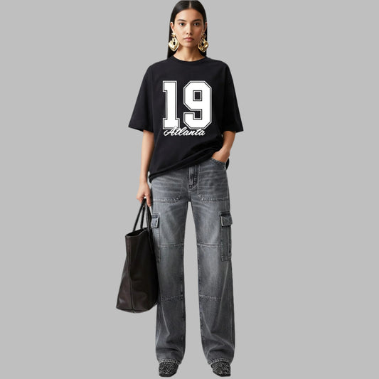 Person wearing a black t-shirt with the number 19 and text, paired with gray cargo pants and holding a black bag on a light gray background.