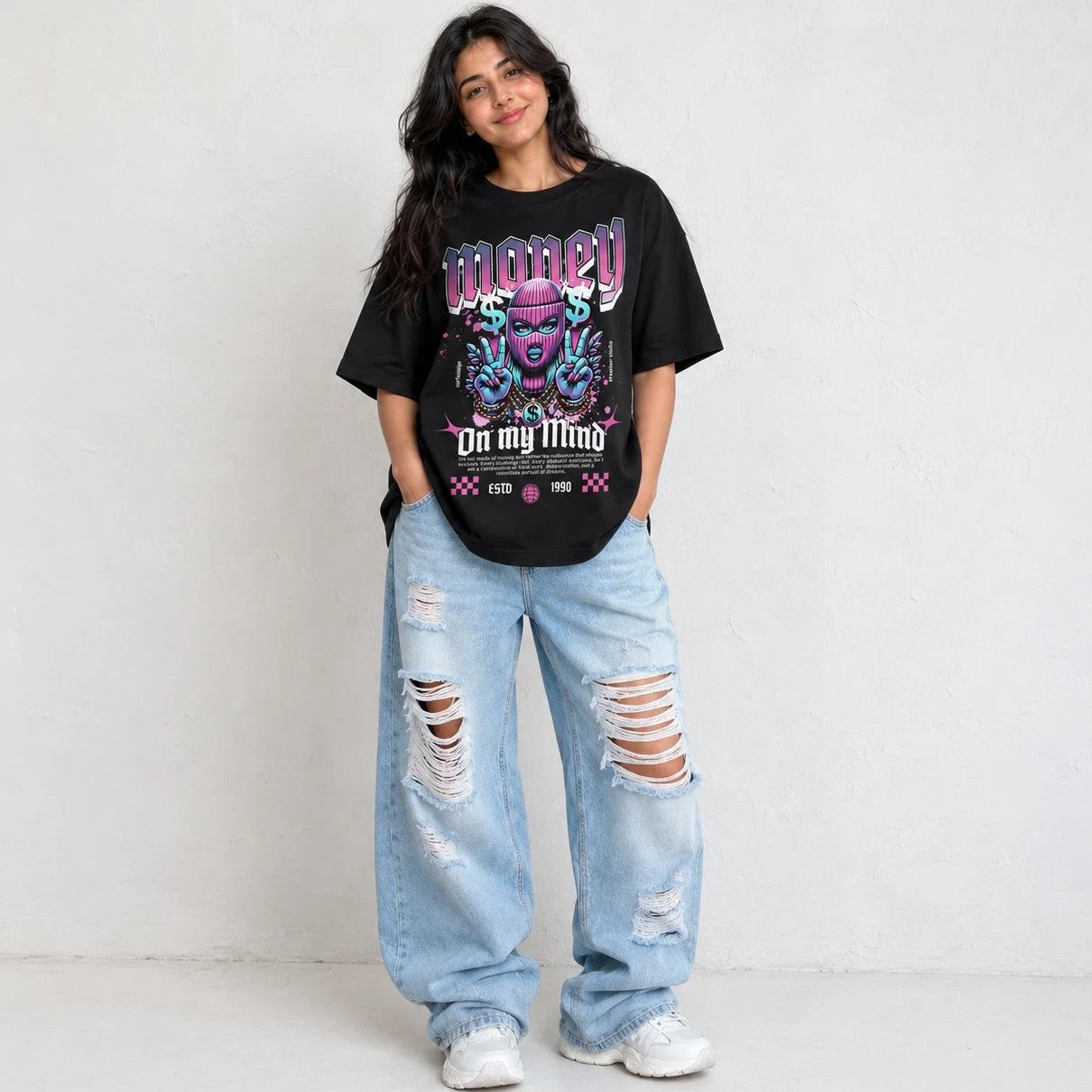 A woman modeling an oversized black graphic t-shirt with a colorful purple and blue design that reads 'MONEY On my Mind', paired with distressed light-wash jeans and white sneakers, standing against a textured off-white wall.