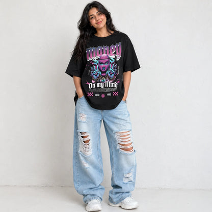 A woman modeling an oversized black graphic t-shirt with a colorful purple and blue design that reads 'MONEY On my Mind', paired with distressed light-wash jeans and white sneakers, standing against a textured off-white wall.