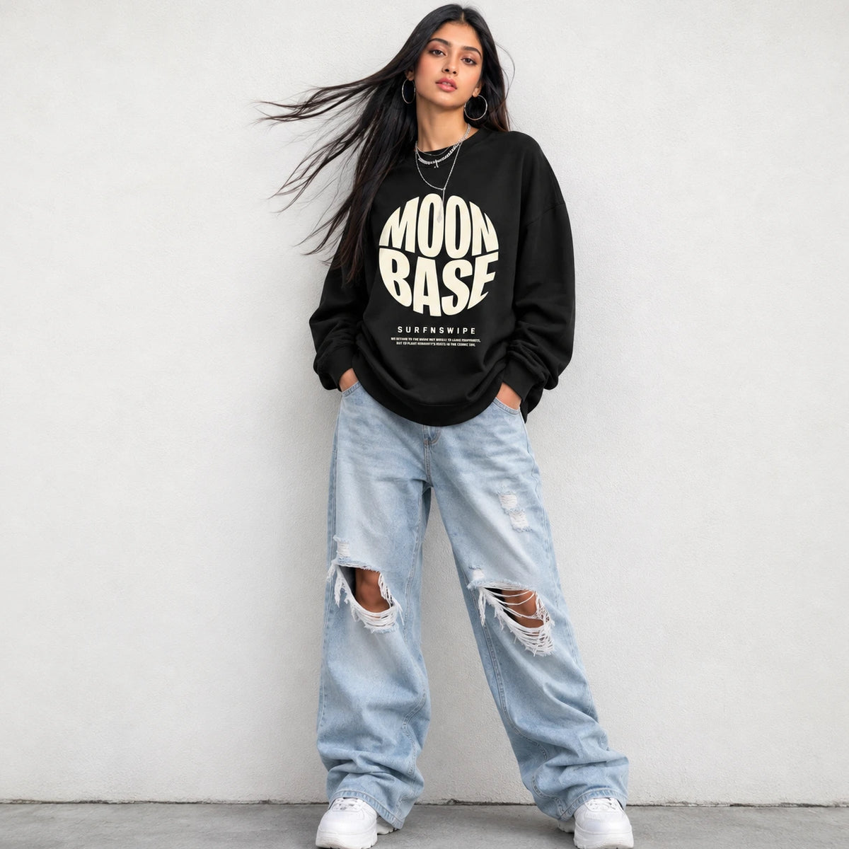 A woman stands against a light gray wall wearing a black sweatshirt with a large cream 'MOON BASE' graphic and distressed light-wash jeans with ripped knees.