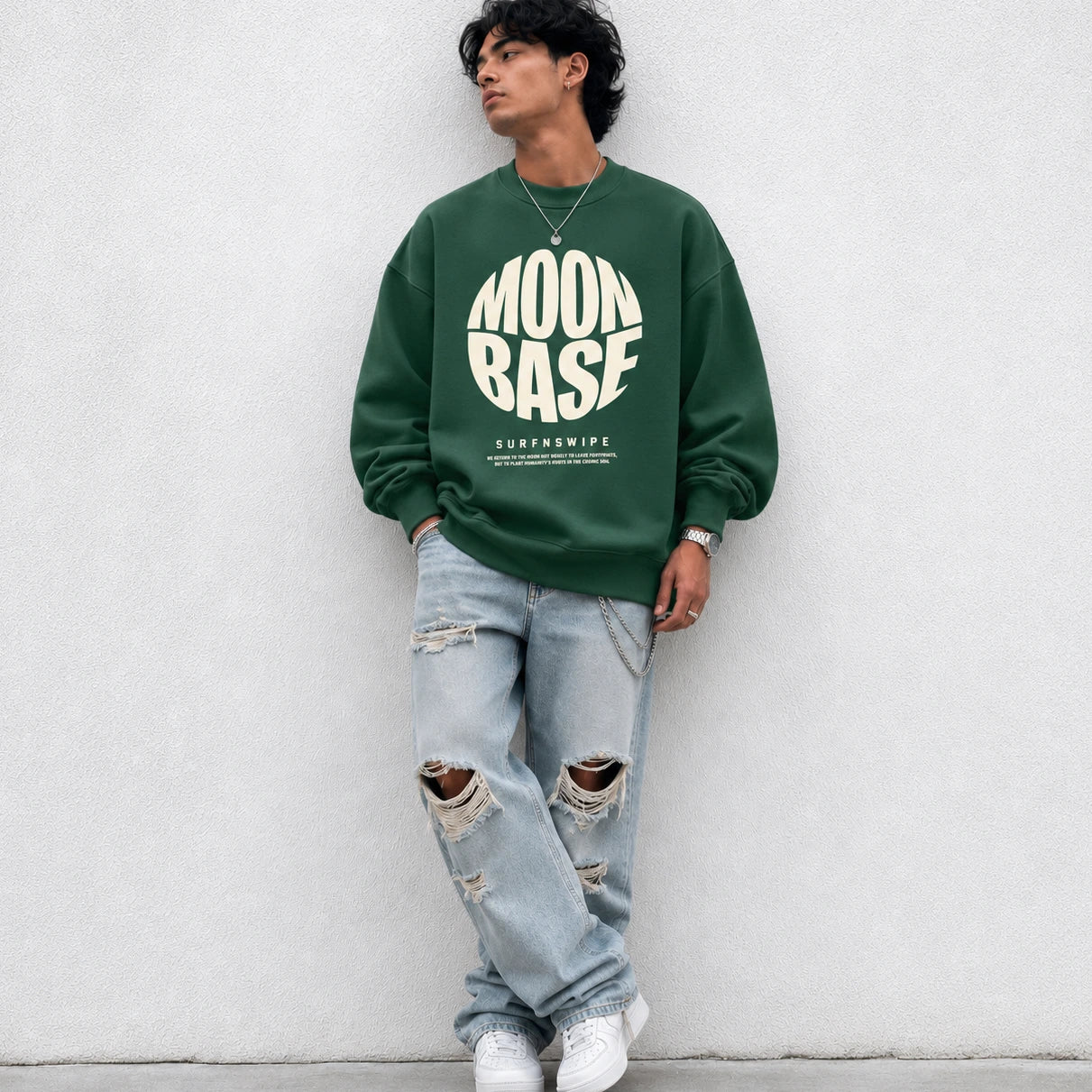 A man wearing a dark green crewneck sweatshirt with a large white 'MOON BASE SURFNSWIPE' graphic on the front, paired with ripped light-wash jeans and white sneakers, leaning against a light gray concrete wall.