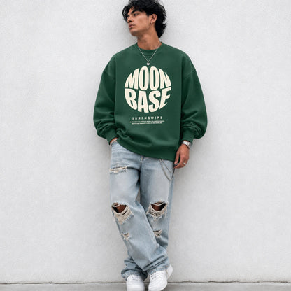 A man wearing a dark green crewneck sweatshirt with a large white 'MOON BASE SURFNSWIPE' graphic on the front, paired with ripped light-wash jeans and white sneakers, leaning against a light gray concrete wall.