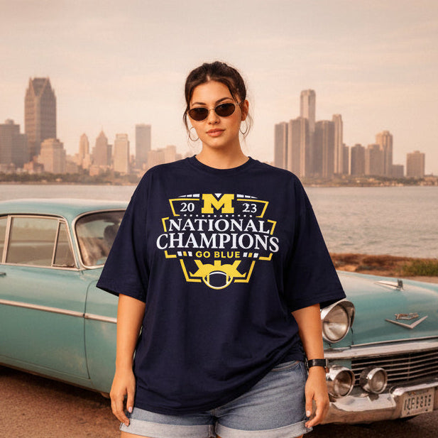 Person wearing a navy blue t-shirt with '2023 National Champions Go Blue' text and graphics, standing in front of a vintage car and city skyline.