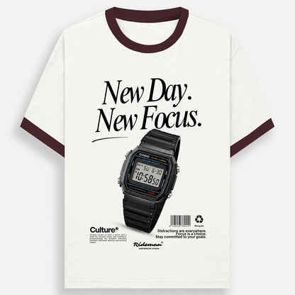 An off-white ringer t-shirt with dark brown trim on the collar and sleeve cuffs, featuring a graphic print of a black Casio watch and the text 'New Day. New Focus.