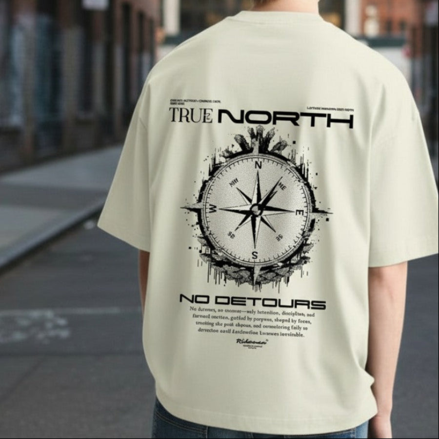 Person wearing a beige t-shirt with 'True North' design on the back, standing on a city street.