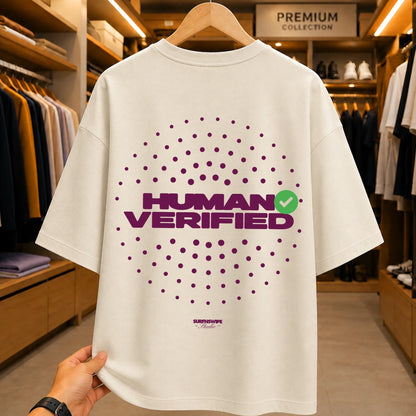 A cream-colored oversized t-shirt with a purple 'HUMAN VERIFIED' graphic and a green checkmark, held up by a hand inside a clothing store.