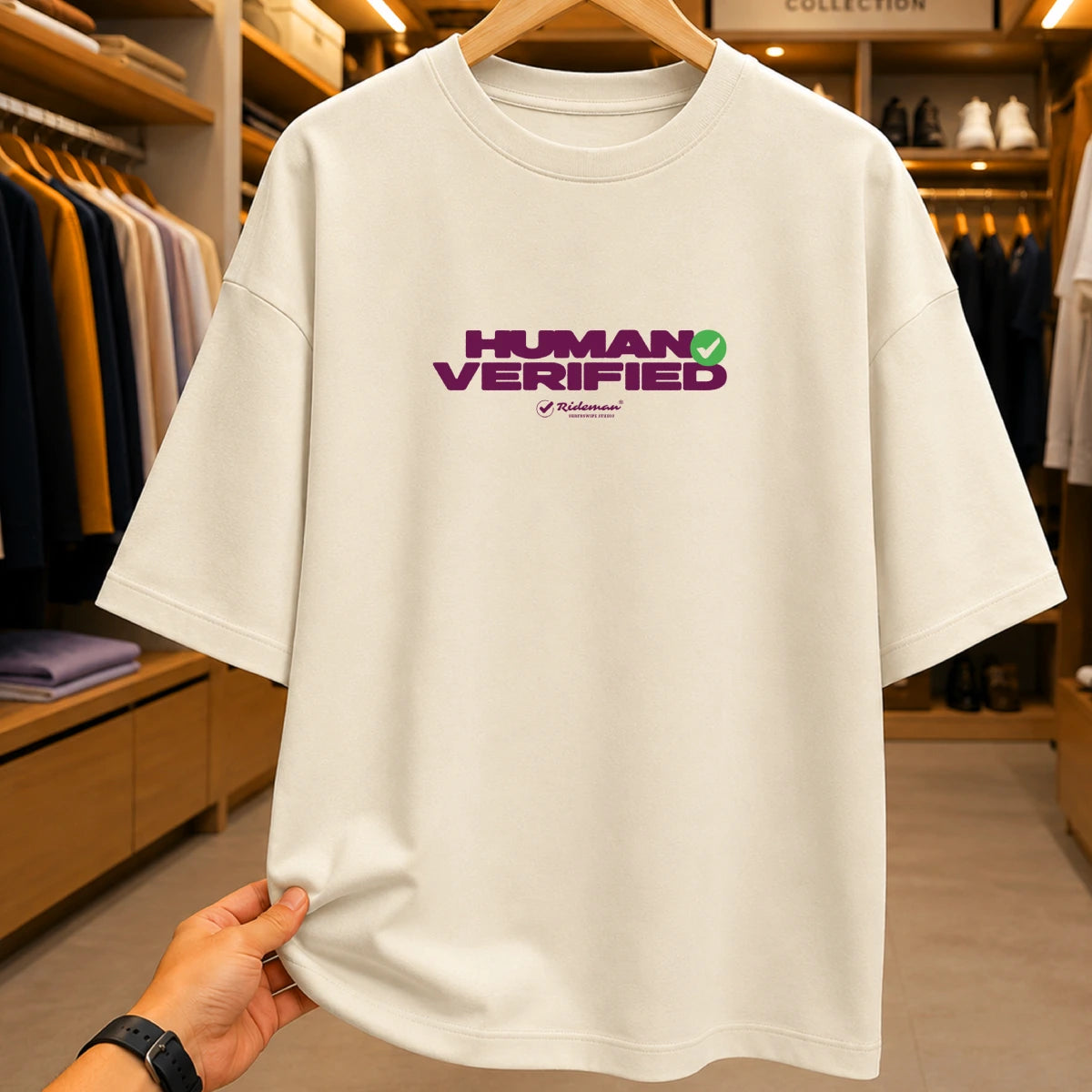 A cream-colored oversized t-shirt with a purple and green 'HUMAN VERIFIED' graphic on the chest, held up by a hand on a wooden hanger inside a clothing store.