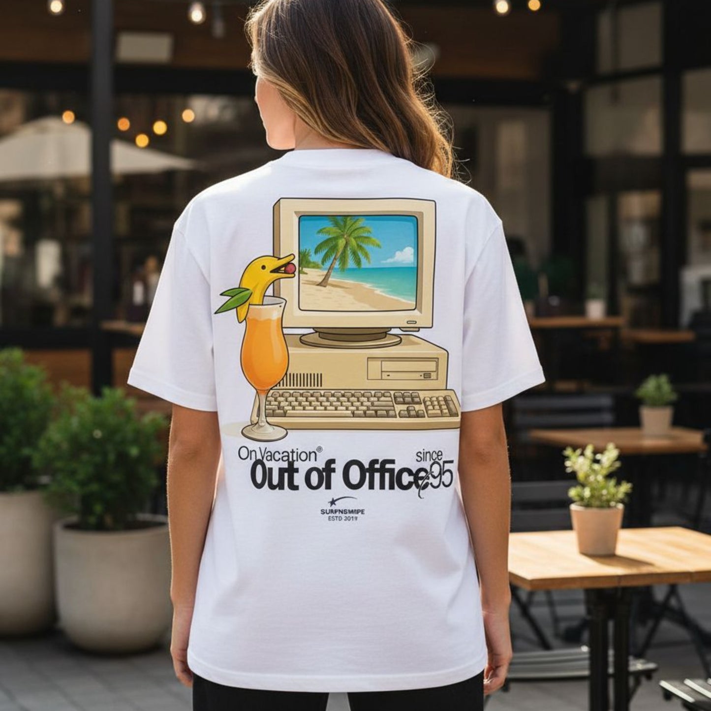 Person wearing a white t-shirt with a graphic design of a computer and beach scene, standing in an outdoor setting.