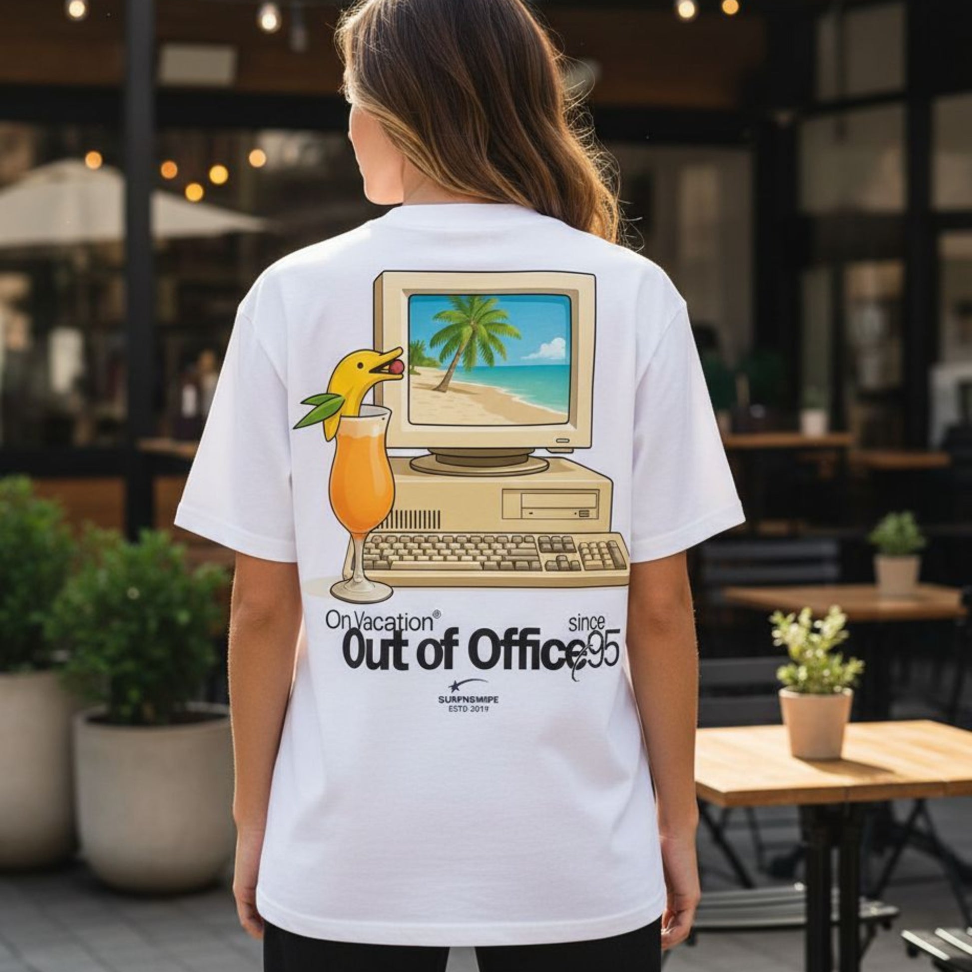 Person wearing a white t-shirt with a graphic design of a computer and beach scene, standing in an outdoor setting.