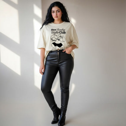 Woman wearing a beige oversized t-shirt with graphic design and black leather pants against a neutral background
