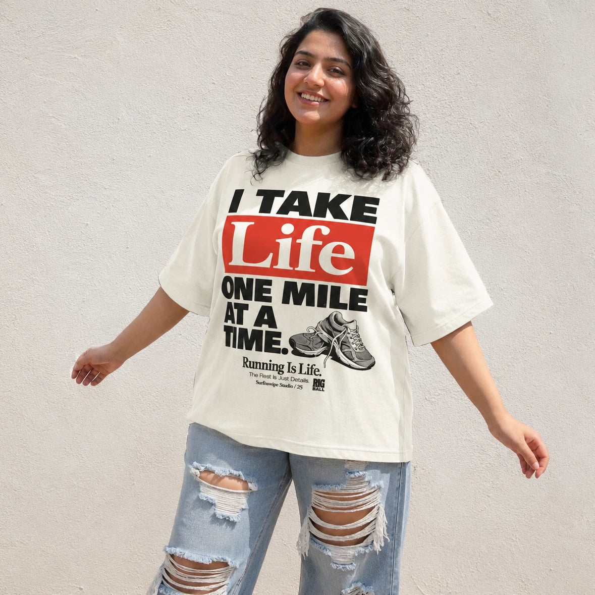 A woman wearing an oversized off-white graphic t-shirt with the text 'I TAKE Life ONE MILE AT A TIME.' in black and red, paired with distressed light blue jeans, standing against a textured off-white wall.