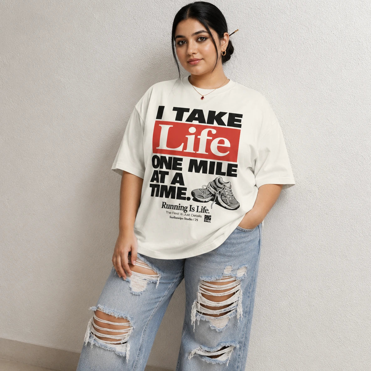 A woman modeling an oversized off-white graphic t-shirt with the text 'I TAKE Life ONE MILE AT A TIME.' in black and red, paired with distressed light blue jeans against a textured light gray wall.