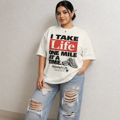 A woman modeling an oversized off-white graphic t-shirt with the text 'I TAKE Life ONE MILE AT A TIME.' in black and red, paired with distressed light blue jeans against a textured light gray wall.
