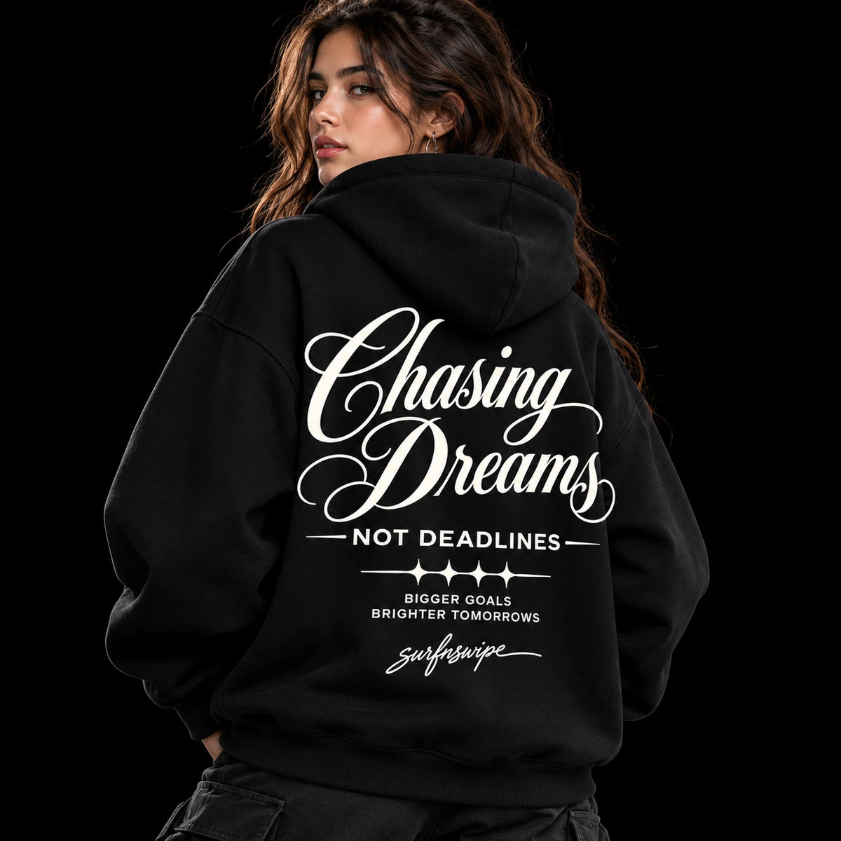 Woman wearing a black Chasing Dreams oversized hoodie with white “Not Deadlines” back lettering and star graphics.

