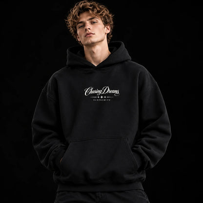  Man wearing a black Chasing Dreams oversized hoodie with a small white chest graphic and kangaroo pocket.
