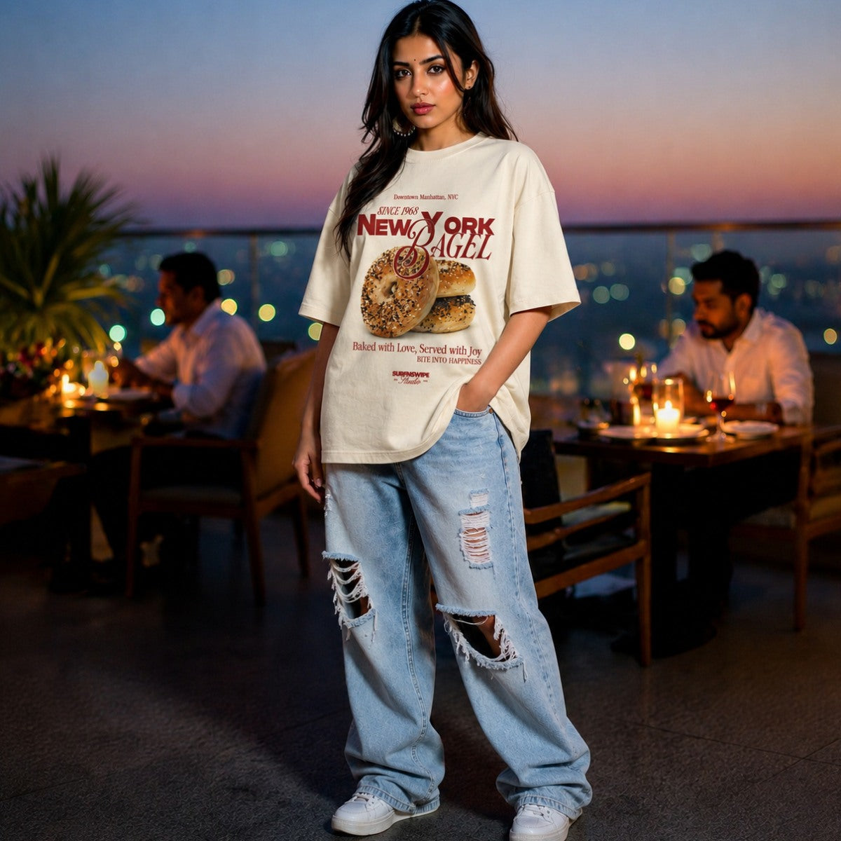 Woman wearing a New York Bagel t-shirt and ripped jeans standing in an outdoor restaurant setting.