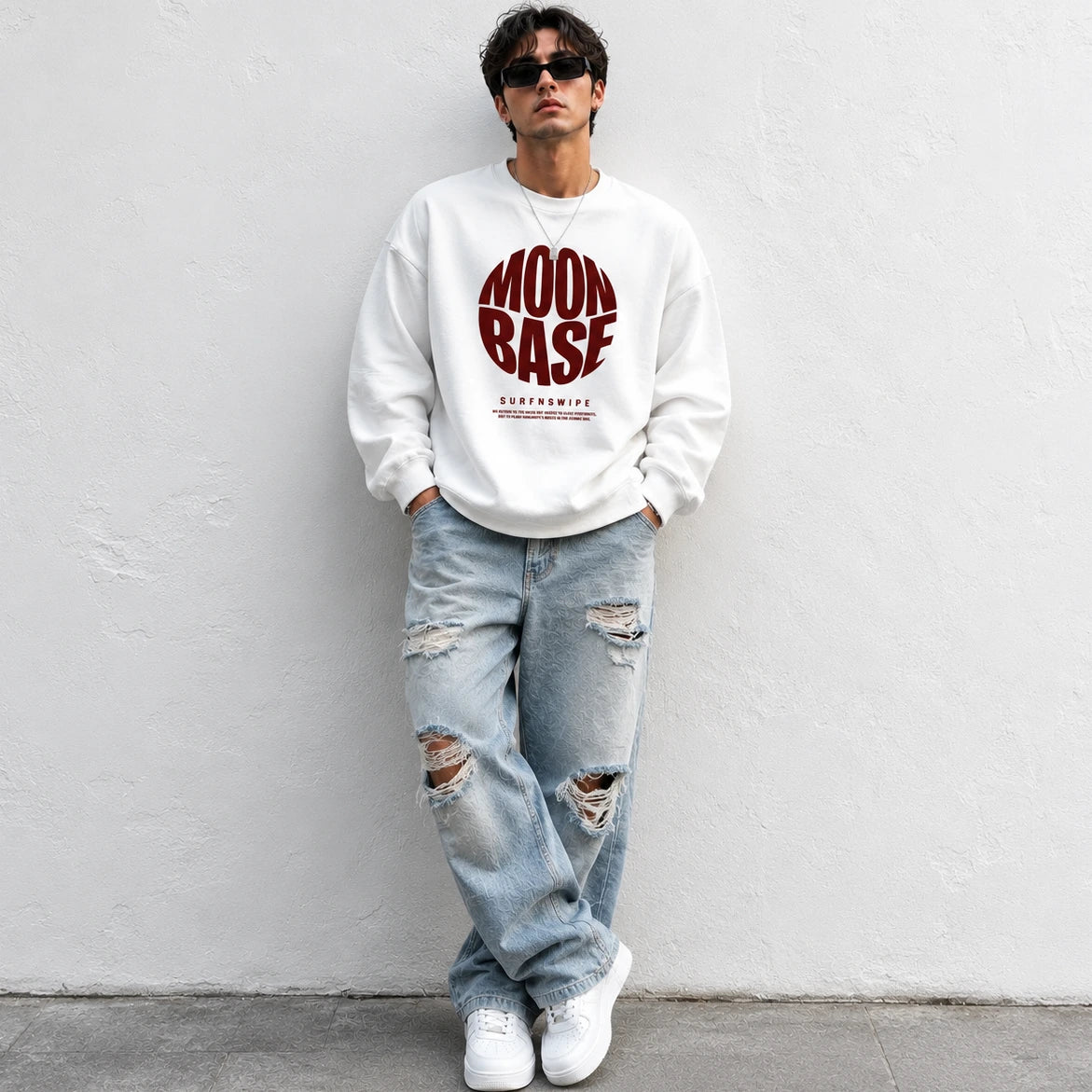 A man wearing a white crewneck sweatshirt with a red 'MOON BASE' graphic, light-wash distressed jeans, and white sneakers, standing against a white textured wall.
