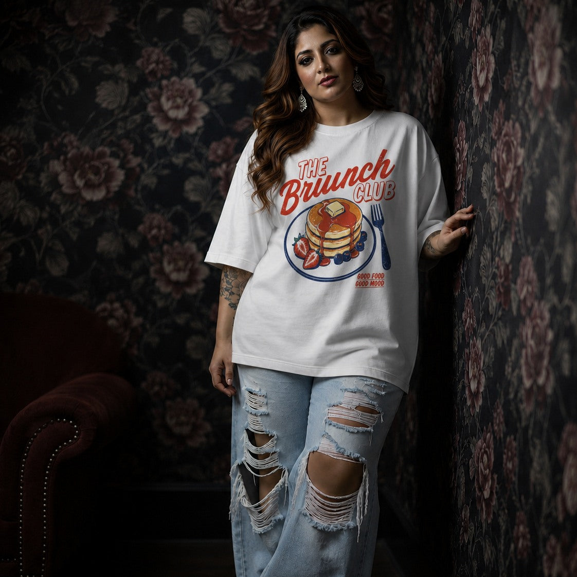 Woman wearing a 'The Brunch Club' t-shirt with a graphic design against a floral-patterned wall.