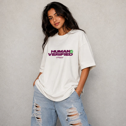 A woman wearing an oversized white t-shirt with a purple and green 'HUMAN VERIFIED' graphic on the chest, paired with light-wash distressed jeans, standing against a textured gray wall.