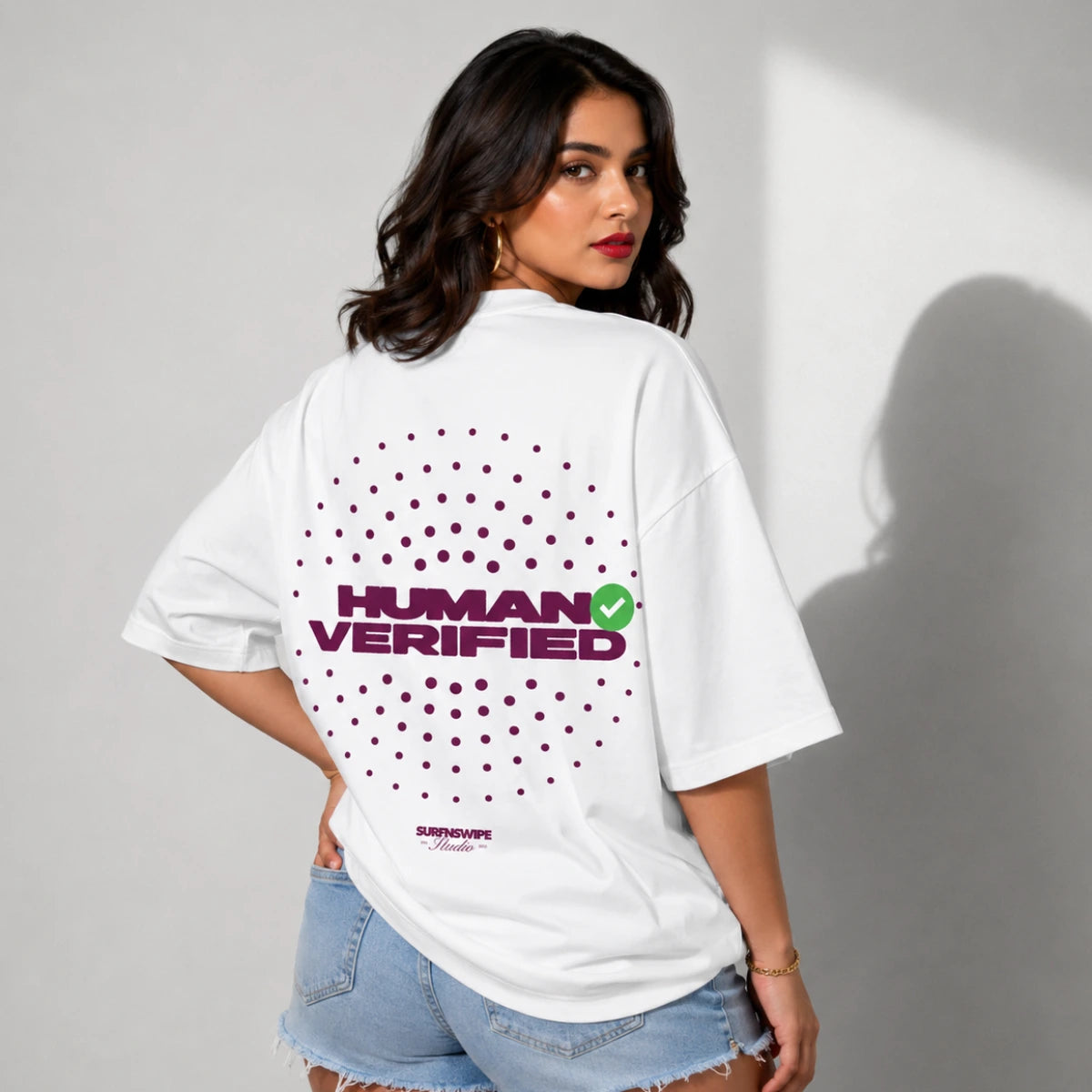 A woman wearing a white oversized t-shirt with a 'HUMAN VERIFIED' graphic on the back, paired with light-wash denim shorts, standing against a neutral gray wall with a soft shadow.