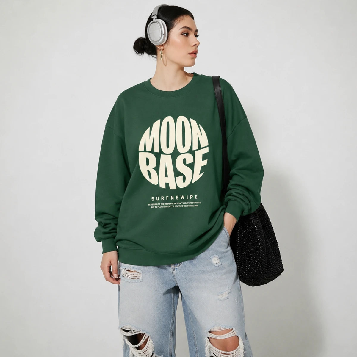 A woman wearing a dark green crewneck sweatshirt with a bold cream graphic that reads 'MOON BASE SURFNSWIPE' on the front, paired with light-wash ripped jeans, silver headphones, and a black woven shoulder bag, set against a plain light gray background.