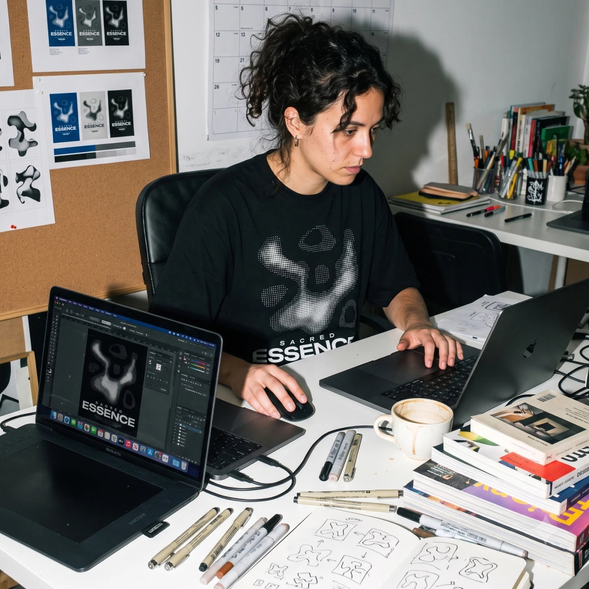 A woman sits at a desk in a creative office workspace, wearing a black t-shirt with a white graphic print that reads 'ESSENCE'. She is surrounded by laptops, sketchbooks, and art supplies on a white desk.
