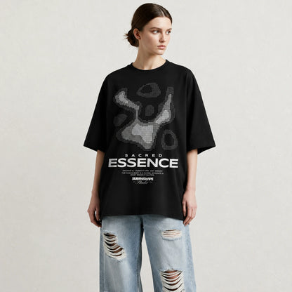 A woman modeling an oversized black t-shirt with a gray abstract graphic and white 'SACRED ESSENCE' text, paired with light-wash distressed jeans against a textured off-white background.