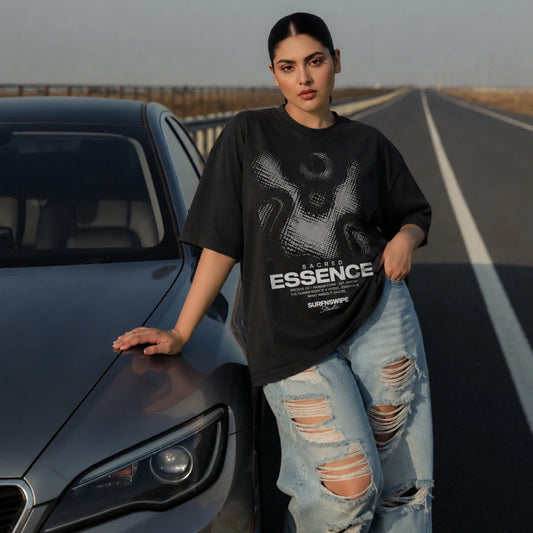 A woman stands on an open road next to a dark gray car, wearing an oversized black graphic T-shirt with the text 'Sacred Essence' and distressed light blue jeans.