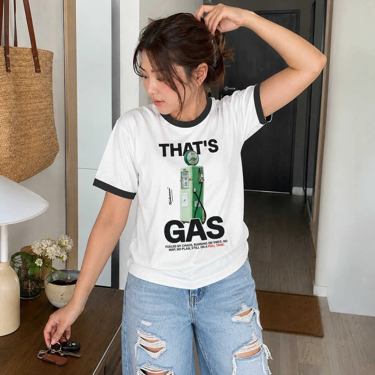 A woman wearing a white ringer T-shirt with black trim and a green graphic print that reads 'THAT'S GAS', paired with distressed light blue jeans, standing in a modern indoor setting.