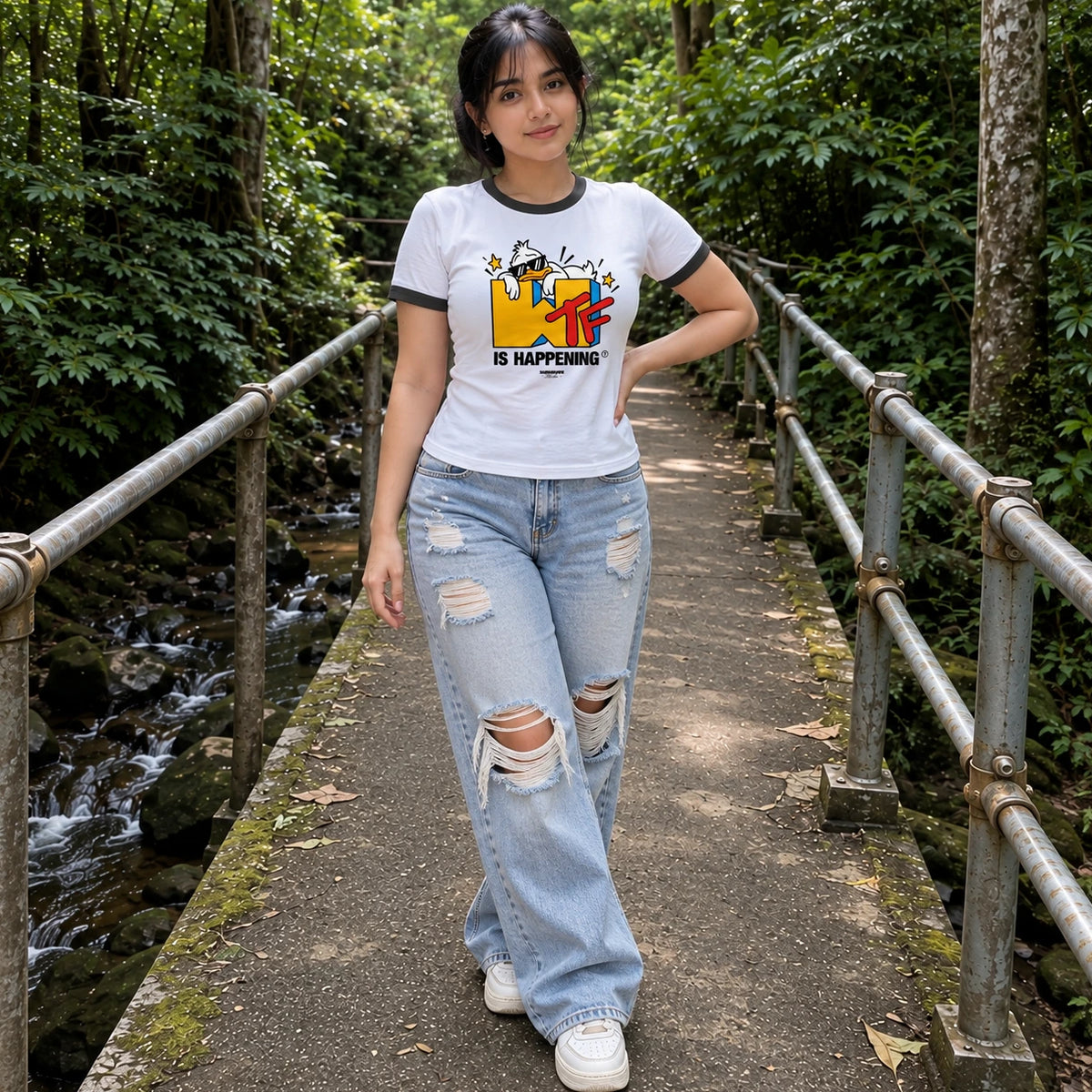 A woman stands on a narrow bridge in a forest wearing a white ringer T-shirt with a colorful graphic and the text 'IS HAPPENING', light-wash distressed jeans, and white sneakers.