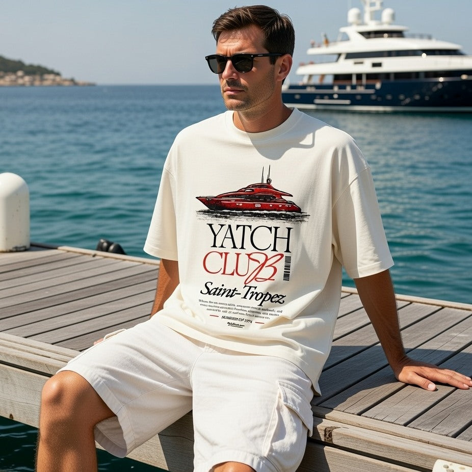 Man wearing a 'Yatch Club Saint-Tropez' t-shirt sitting on a dock with a yacht in the background.